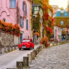 O stradă primitoare din Montmartre, cu frunze de toamnă care acoperă aleile pietruite. O mașină clasică roșie Diane se evidențiază pe fundalul pitoresc al cartierului parizian.