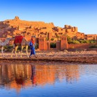 Satul fortificat Ait Benhaddou, patrimoniu UNESCO din Maroc, se reflectă în apă printre cămile, ziduri din pământ și lumina apusului.