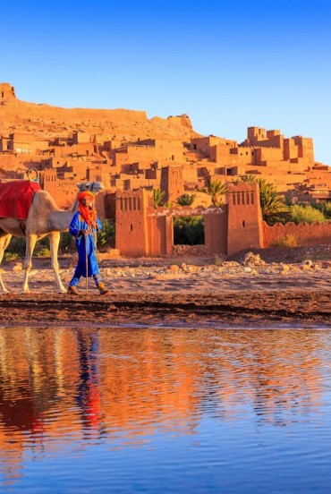 Satul fortificat Ait Benhaddou, patrimoniu UNESCO din Maroc, se reflectă în apă printre cămile, ziduri din pământ și lumina apusului.
