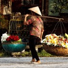 Un vânzător vietnamez în Hanoi, printre străzi istorice și piețe, cu coșuri cu flori care aduc parfumuri și culori în viața cotidiană a orașului.