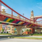 Un pod colorat din Hanoi, cu arhitectură europeană și tonuri vii de curcubeu, se ridică printre clădiri moderne și un cer albastru, într-o scenă plină de lumină și viață.