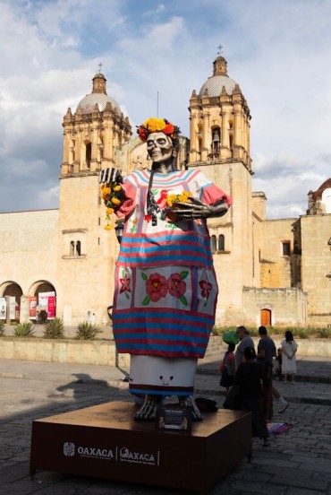 Catrină colorată în fața Bisericii Santo Domingo din Oaxaca: figura de Día de los Muertos se evidențiază pe fundalul arhitecturii baroce, în timp ce vizitatorii traversează piața