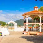 Piața din Zinacantán, în fața Bisericii San Lorenzo, prezintă un foișor elegant alb și portocaliu, decorat cu stegulețe colorate, înconjurat de coline verzi și viață locală autentică.