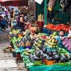 O femeie și o fetiță tzotzil în piața din San Cristóbal expun fructe proaspete pe țesături verzi și bleu, creând o scenă autentică plină de culoare, tradiție și viață indigenă cotidiană.