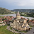 Vedere aeriană a impunătoarei Catedrale Svetitskhoveli din Mtskheta, vechi centru religios al Georgiei, cu râul Mtkvari și dealurile verzi ce înconjoară orașul.