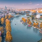 Vedere aeriană a orașului Tbilisi, cu râul Mtkvari (Kura) traversând orașul, Podul Păcii și parcurile de pe mal ce îmbină arhitectura istorică și modernă.