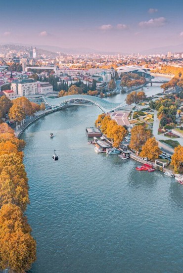 Vedere aeriană a orașului Tbilisi, cu râul Mtkvari (Kura) traversând orașul, Podul Păcii și parcurile de pe mal ce îmbină arhitectura istorică și modernă.
