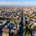 Vedere aeriană a Erevanului cu animatul bulevard Mashtots care traversează orașul printre clădiri, alei cu copaci și cartiere istorice, cu impunătorul Munte Ararat la orizont.