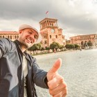 Turist zâmbitor făcând un selfie în fața fântânilor din Piața Republicii din Erevan. În fundal se vede turnul cu ceas al clădirii Guvernului și arhitectura elegantă armeană.