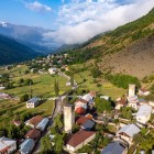 Vedere aeriană a orașului Mestia, în inima Svaneti, cu turnuri medievale din piatră printre pajiști verzi și peisaje alpine, dominate de vârfurile impunătoare ale Caucazului.