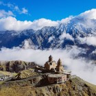 Priveliște spectaculoasă asupra Bisericii Sfânta Treime din Gergeti, cocoțată între Munții Caucaz cu impunătorul Munte Kazbek în fundal și nori care învăluie valea.