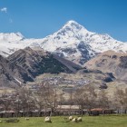 Priveliște asupra satului Gergeti la poalele Muntelui Kazbek, cu case adunate în vale și vârfurile înzăpezite ale Marelui Caucaz dominând orizontul.
