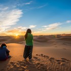 Răsărit printre dunele din Merzouga, cu două călătoare care privesc soarele ridicându-se peste Sahara, între liniște, nisip auriu și orizonturi infinite.