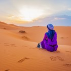 Călătoare așezată pe dunele din Merzouga, privind răsăritul peste Sahara, între liniște, nisip auriu și panorame infinite ale deșertului.