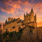 Silueta de basm a Alcázarului din Segovia domină promontoriul stâncos, în timp ce cerul la apus îmbracă turnurile, crenelurile și zidurile într-o lumină aurie ce evidențiază eleganța arhitecturii sale medievale.