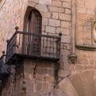 Un balcon din fier forjat se desprinde din fațada de piatră a centrului istoric din Cáceres, lângă o stemă nobiliară ce evocă trecutul aristocratic al orașului medieval.