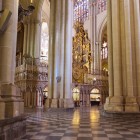 Pilonii gotici impunători delimitează navele Catedralei din Toledo; pardoseala geometrică îndrumă privirea spre retablul aurit, încadrat de arcade și vitralii.