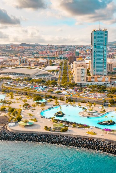Parcul Maritim César Manrique se deschide spre ocean cu piscine cu apă sărată, palmieri și terase pentru plajă, în timp ce Turnurile din Santa Cruz conturează silueta modernă a capitalei insulei Tenerife.