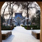 Porțile din lemn sculptat încadrează drumul spre Madrasa Nadir Divan-Begi, dezvăluind călătorului primele detalii ale faianței și atmosferei istorice din Buhara.